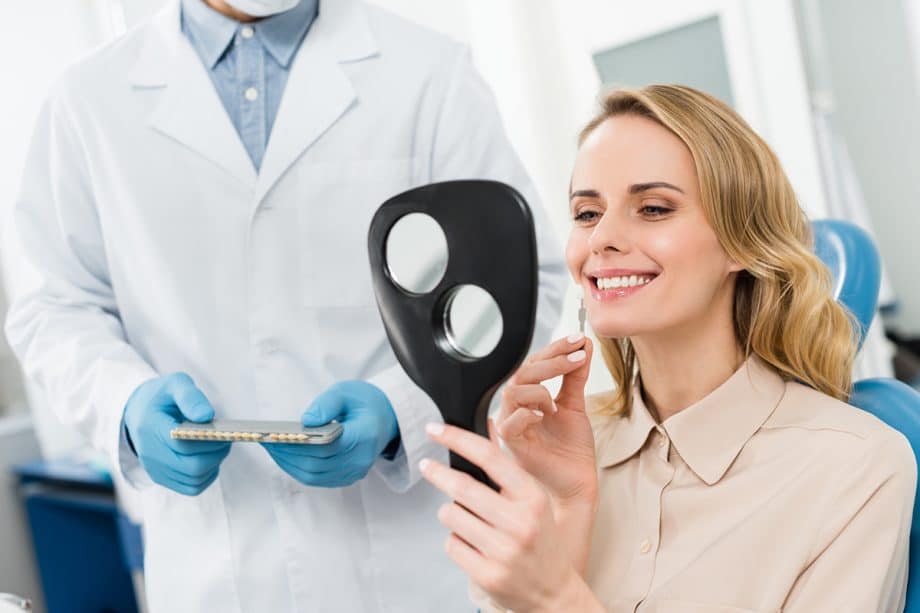 a woman looking at her dental veneers in the mirror in a dental chair with a dentist standing by her side.