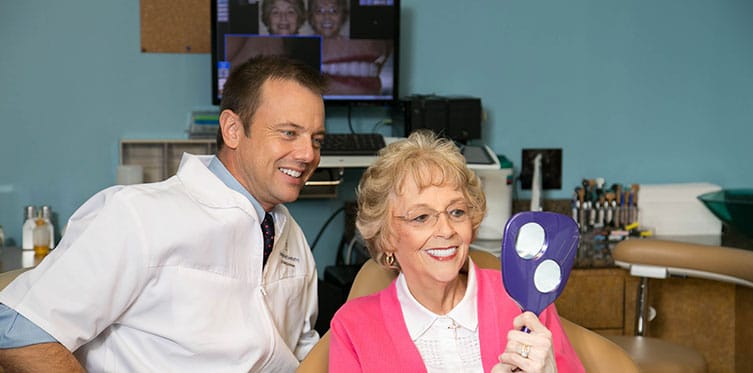 older woman in dental chair holding hand mirror, smiling with dentist beside her